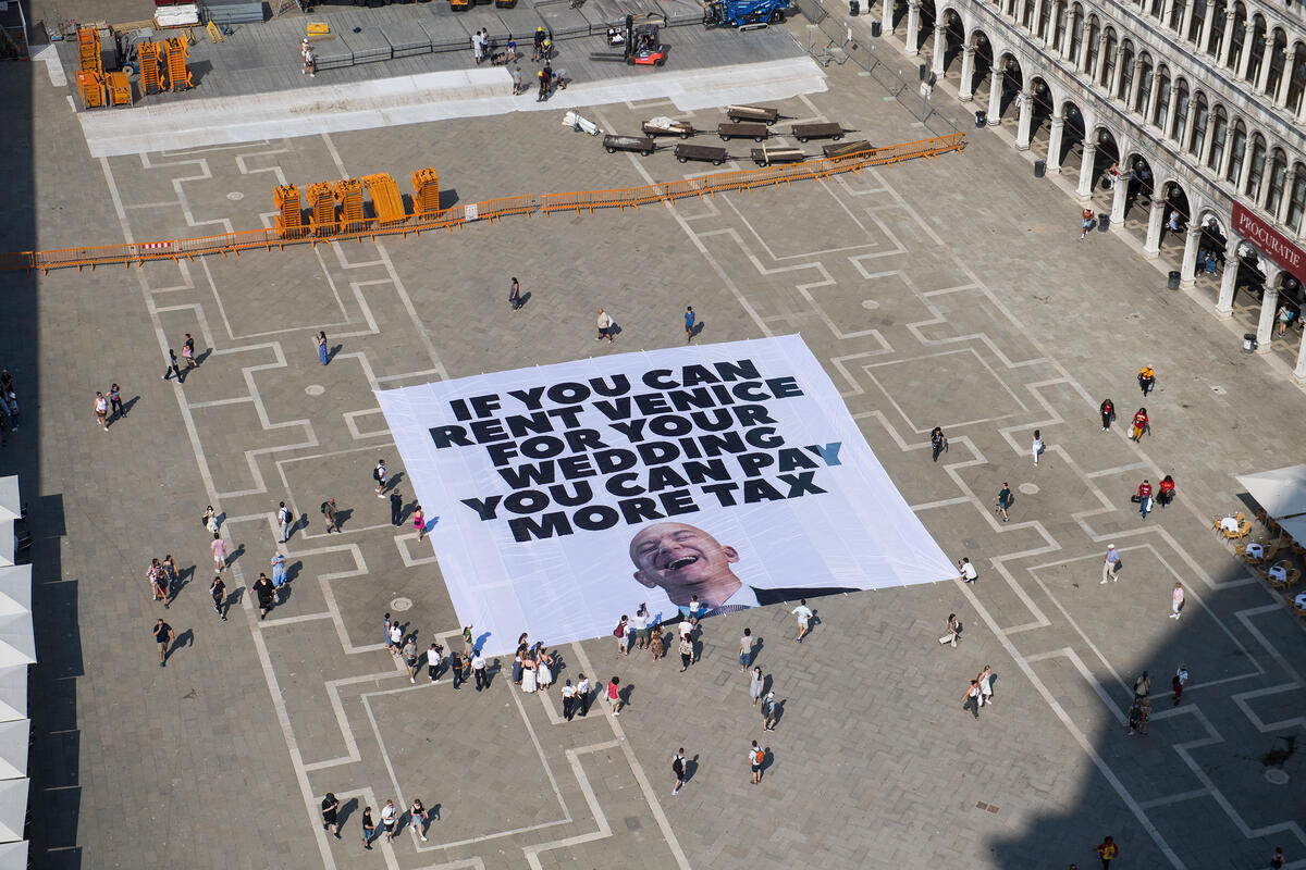  L'immagine mostra una piazza San Marco con un grande striscione bianco al centro. Il testo sullo striscione recita: "IF YOU CAN RENT VENICE FOR WEDDING YOU CAN PAY MORE TAX". Sotto il testo, c'è un'immagine di un uomo sorridente. La piazza è pavimentata con un disegno geometrico e ci sono persone che camminano o si riuniscono intorno allo striscione. Sullo sfondo, si vedono edifici storici e alcune strutture di costruzione. L'atmosfera sembra essere quella di una protesta o di un evento pubblico.