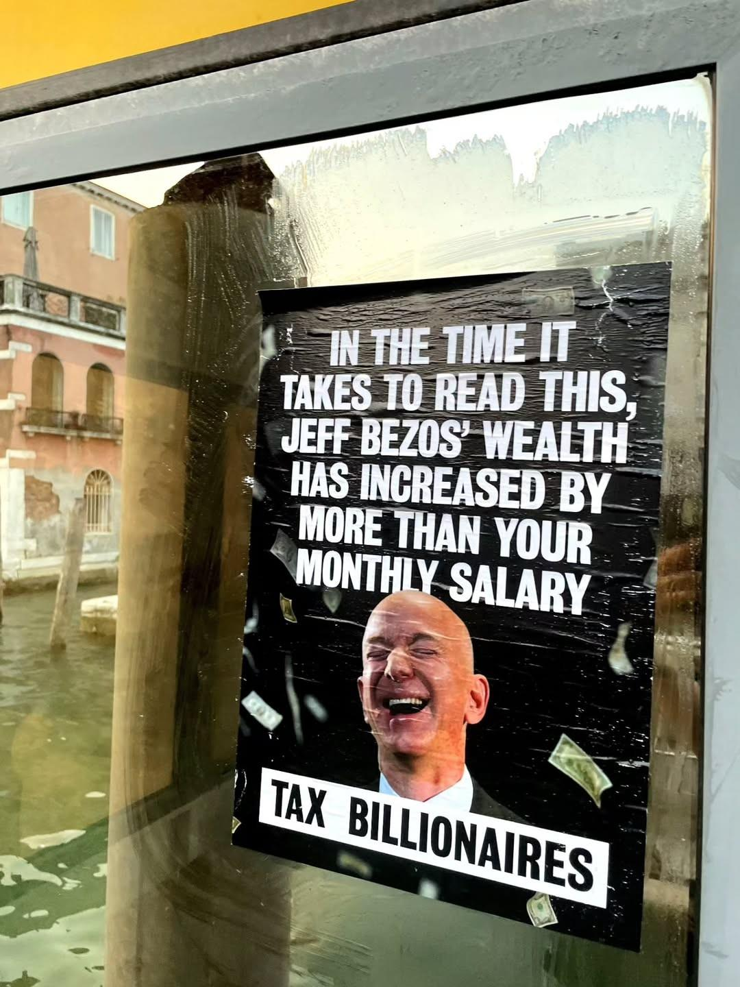L'immagine mostra un poster attaccato a una finestra, con un sfondo che riflette un canale e edifici tipici di Venezia. Il poster è nero con testo bianco e presenta un'immagine di Jeff Bezos, con un'espressione di gioia, circondato da banconote che sembrano volare. Il testo recita: "IN THE TIME IT TAKES TO READ THIS, JEFF BEZOS' WEALTH HAS INCREASED BY MORE THAN YOUR MONTHLY SALARY" e "TAX BILLIONAIRES". Questo poster sembra essere un messaggio politico che critica la ricchezza dei miliardari e la loro crescita economica rapida.